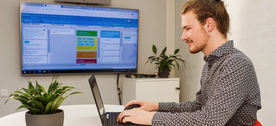 Ein Mann sitzt an einem modernen Schreibtisch und bearbeitet etwas am Laptop. Im Hintergrund ist ein Kanban-Board auf einem Bildschirm zu sehen. Foto: Thomas Grimm