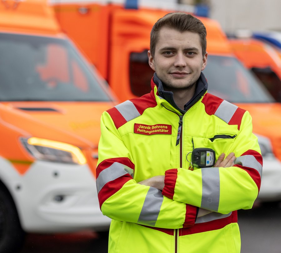 Ein Rettungssanitäter steht in Dienstkleidung vor Fahrzeugen des Rettungsdienstes